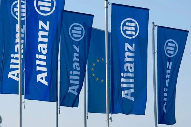 Row of blue Allianz flags with white logos and text flying in the wind, with an EU flag visible among them against a light sky