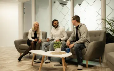 Three people having a casual meeting in a modern office lounge area, sitting on grey couches around a small round table