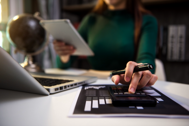 Close-up of hands using a calculator while reviewing documents on a laptop