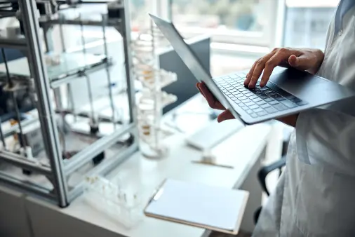 Person typing on a laptop keyboard in a laboratory setting with test tubes and scientific equipment visible on the desk