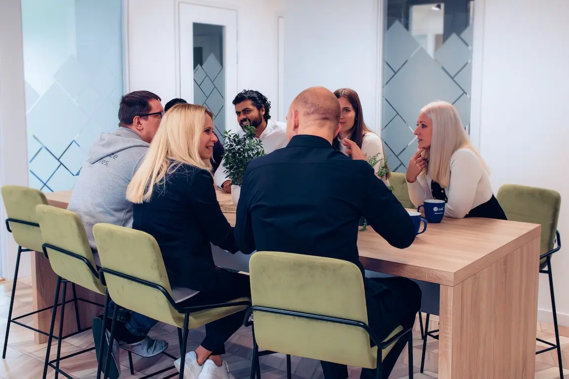 A group of employees having an informal meeting around a wooden table, sitting on yellow chairs in a modern office space