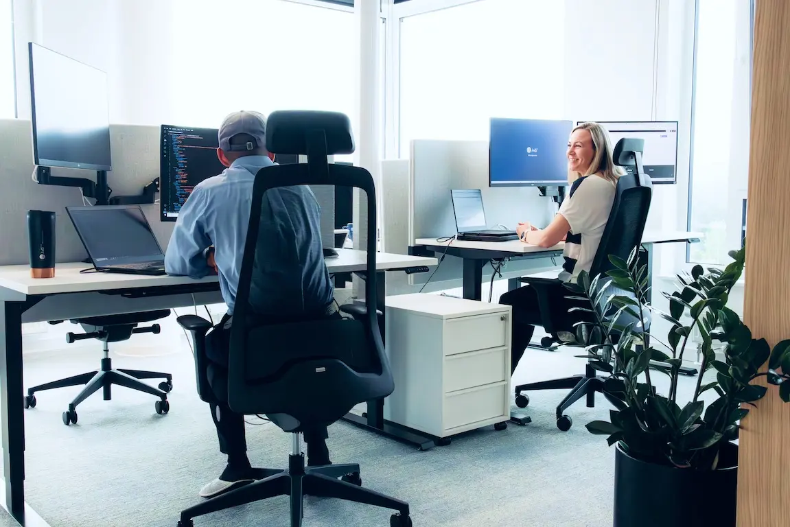 Two colleagues having a walking conversation in a bright office hallway with white walls and potted plants