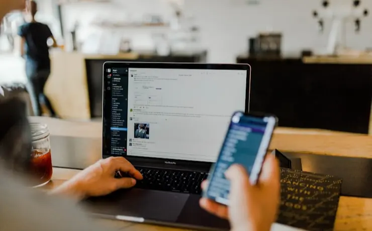 Person using laptop and smartphone simultaneously at a wooden desk with a drink nearby