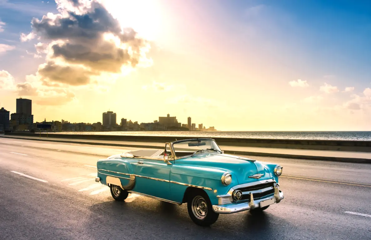 Vintage turquoise Chevrolet convertible on a waterfront street at sunset with a city skyline in the background