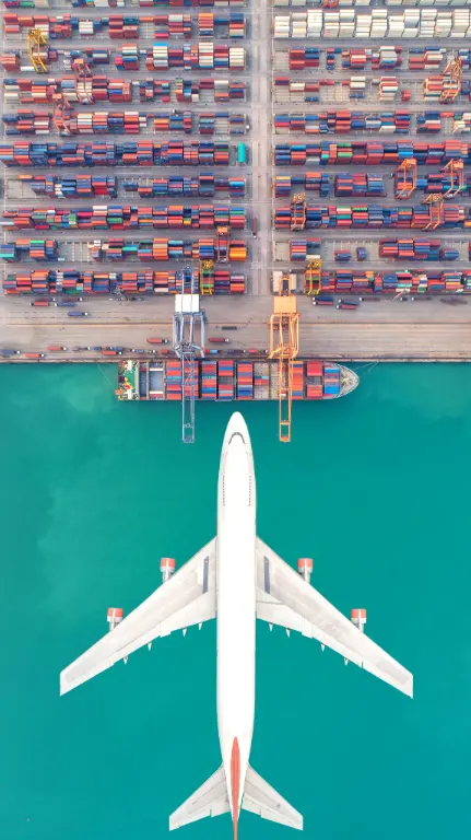 Aerial view of a white cargo plane over turquoise water approaching a container port with stacked shipping containers and cargo ships