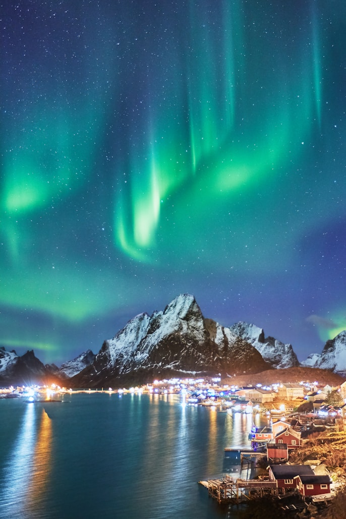 Northern lights (aurora borealis) illuminating the night sky over a small coastal town with snow-covered mountains in the background