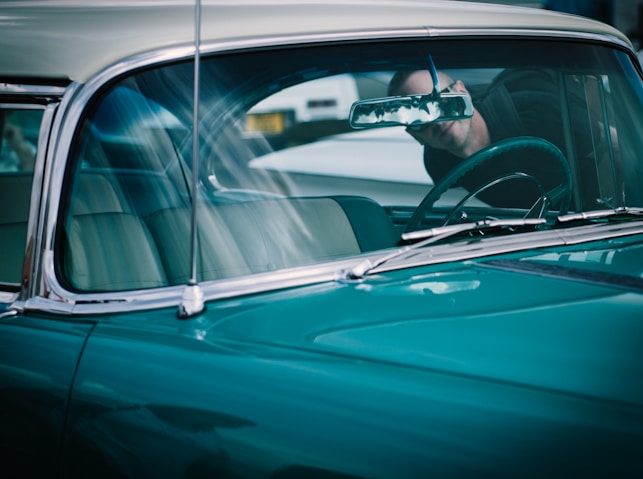 Close-up view of a vintage car's windshield and turquoise hood, with chrome trim and partial view of the steering wheel through the glass